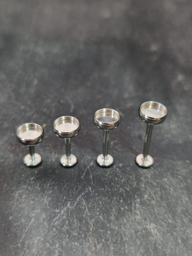 May include: Four silver-colored jewelry posts with a flat, circular top and a long, slender stem. The posts are arranged in a row, with the stems and tops of each post visible. The posts are on a dark, textured surface.