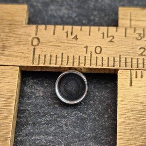 May include: A small, round, silver-colored object with a dark center, positioned near a vintage wooden ruler. The ruler has black markings and numbers. The object is approximately 0.5 inches in diameter.