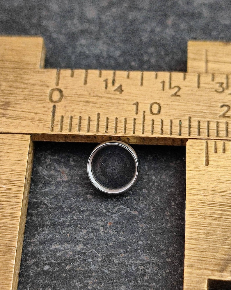 May include: A small, round, silver-colored object with a dark center, positioned near a vintage wooden ruler. The ruler has black markings and numbers. The object is approximately 0.5 inches in diameter.