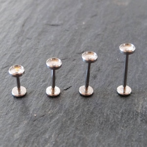 May include: Four silver-colored, labret-style lip or ear studs. Each stud has a flat, round base and a matching top, connected by a straight post. The studs are arranged in a row, varying slightly in post length.
