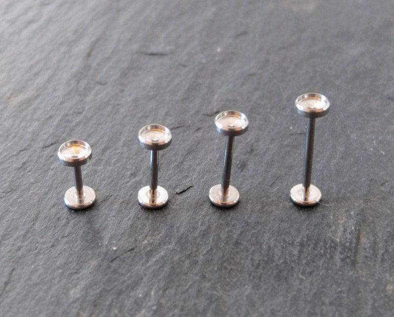 May include: Four silver-colored, labret-style lip or ear studs. Each stud has a flat, round base and a matching top, connected by a straight post. The studs are arranged in a row, varying slightly in post length.