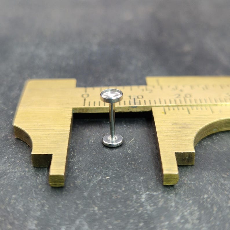 May include: A small, silver-colored labret stud with a flat base and a circular top. The stud is positioned on a brass caliper, with the measurement scale visible. The background is a dark, textured surface.