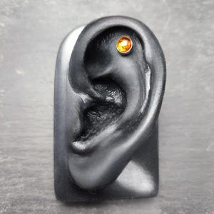 May include: A black ear model with a small, round, amber-colored stud earring. The earring is set in a silver-colored metal. The ear model is displayed on a gray surface.
