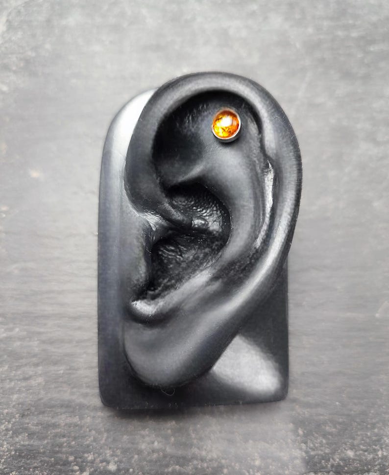 May include: A black ear model with a small, round, amber-colored stud earring. The earring is set in a silver-colored metal. The ear model is displayed on a gray surface.