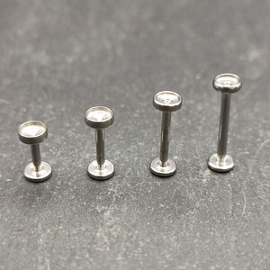May include: Four silver body jewelry pieces with clear gem tops. The pieces are different sizes, with the smallest on the left and the largest on the right.