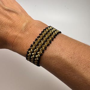 May include: A black and gold beaded bracelet with a geometric pattern. The bracelet is made with small, round beads and is worn on a person's wrist.