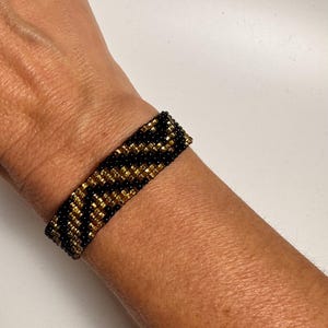 May include: A black and gold beaded bracelet with a geometric pattern. The bracelet is worn on a person's wrist.