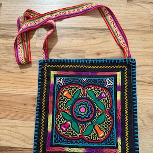 May include: Black square shoulder bag with colorful floral embroidery. The bag features a vibrant design with red, green, orange, and purple accents. The strap is a patterned ribbon with red, green, and purple stripes. The bag is on a wooden surface.