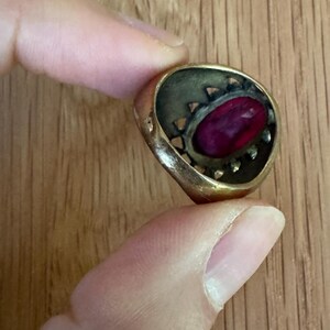 May include: A gold-toned metal ring featuring a central oval-shaped deep magenta stone. The setting has a decorative openwork gallery with small triangular cutouts surrounding the stone, giving the piece a textured and layered appearance.