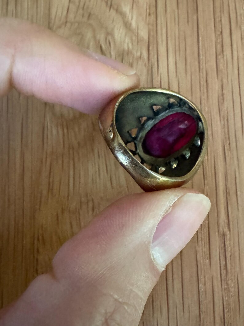 May include: A gold-toned metal ring featuring a central oval-shaped deep magenta stone. The setting has a decorative openwork gallery with small triangular cutouts surrounding the stone, giving the piece a textured and layered appearance.
