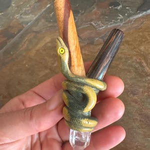 Pode incluir: Uma varinha de madeira esculpida com um desenho de cobra enrolada em verde e amarelo, enrolada em torno de uma ponta de cristal transparente. A varinha tem dois cabos de madeira, um castanho claro e outro castanho escuro.