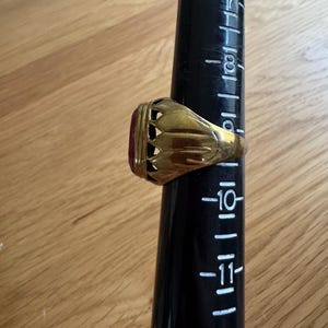 May include: A gold-toned ring featuring a deep red oval stone set in a decorative, scalloped metal gallery. The ring is positioned on a black plastic ring mandrel marked with size numbers, showing the band rests near the size 10.5 mark.
