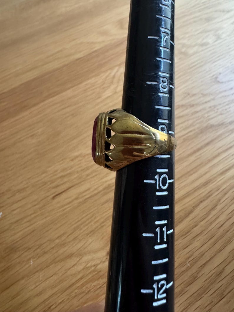 May include: A gold-toned ring featuring a deep red oval stone set in a decorative, scalloped metal gallery. The ring is positioned on a black plastic ring mandrel marked with size numbers, showing the band rests near the size 10.5 mark.