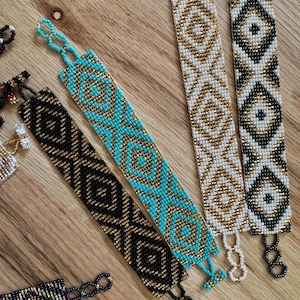 May include: Several beaded bracelets with geometric patterns in various colors, including turquoise, gold, black, and white. The bracelets are laid out on a wooden surface, showcasing their intricate designs and craftsmanship.