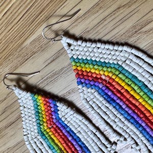 May include: A pair of long, rainbow-coloured beaded earrings with white beads. The earrings are made of small, round beads that are strung together to create a fringe effect. The rainbow colours are arranged in a gradient pattern, starting with red at the top and ending with purple at the bottom.