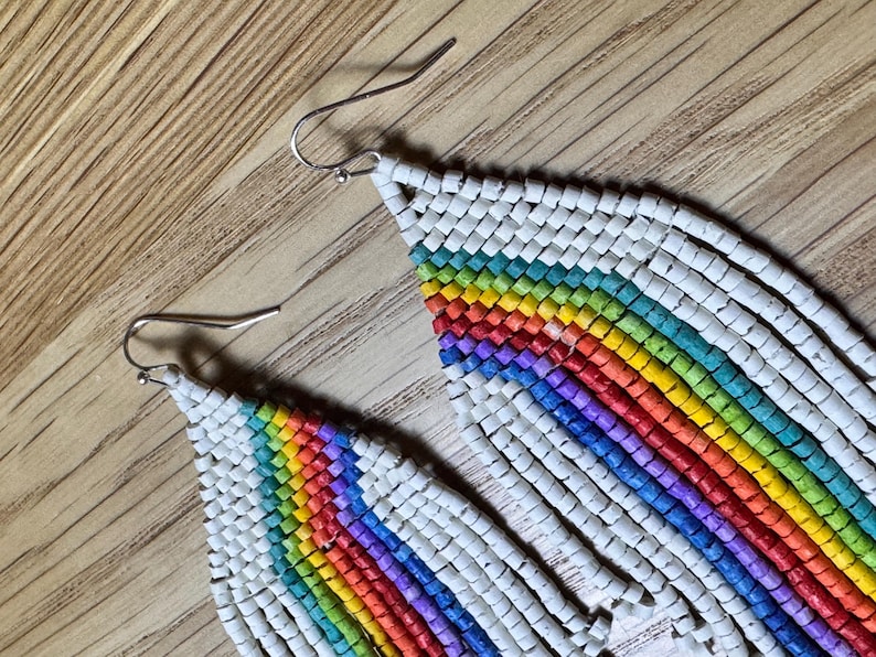 May include: A pair of long, rainbow-coloured beaded earrings with white beads. The earrings are made of small, round beads that are strung together to create a fringe effect. The rainbow colours are arranged in a gradient pattern, starting with red at the top and ending with purple at the bottom.