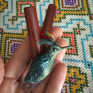 Könnte beinhalten: Ein Haaraccessoire mit zwei dunkel rotbraunen Holzstäben und einem geformten grünen Kolibri mit irisierenden Details. Der Vogel hat einen kleinen Kamm aus roten, gelben und blaugrünen Federn auf dem Kopf sowie eine klare, hellviolette Kristallbasis.