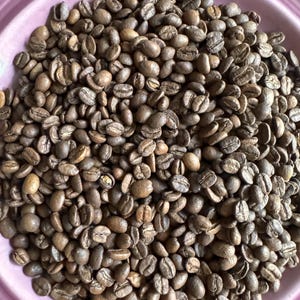 May include: A close-up view of a bowl filled with roasted coffee beans. The beans are a dark brown color and have a slightly oily sheen. The bowl is a light pink color and has a smooth, glossy finish.