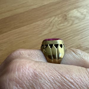 May include: A gold ring featuring a prominent, rectangular, deep pink gemstone set in a metal band. The setting displays a series of triangular cutouts along the gallery, creating a crown-like appearance around the base of the stone.