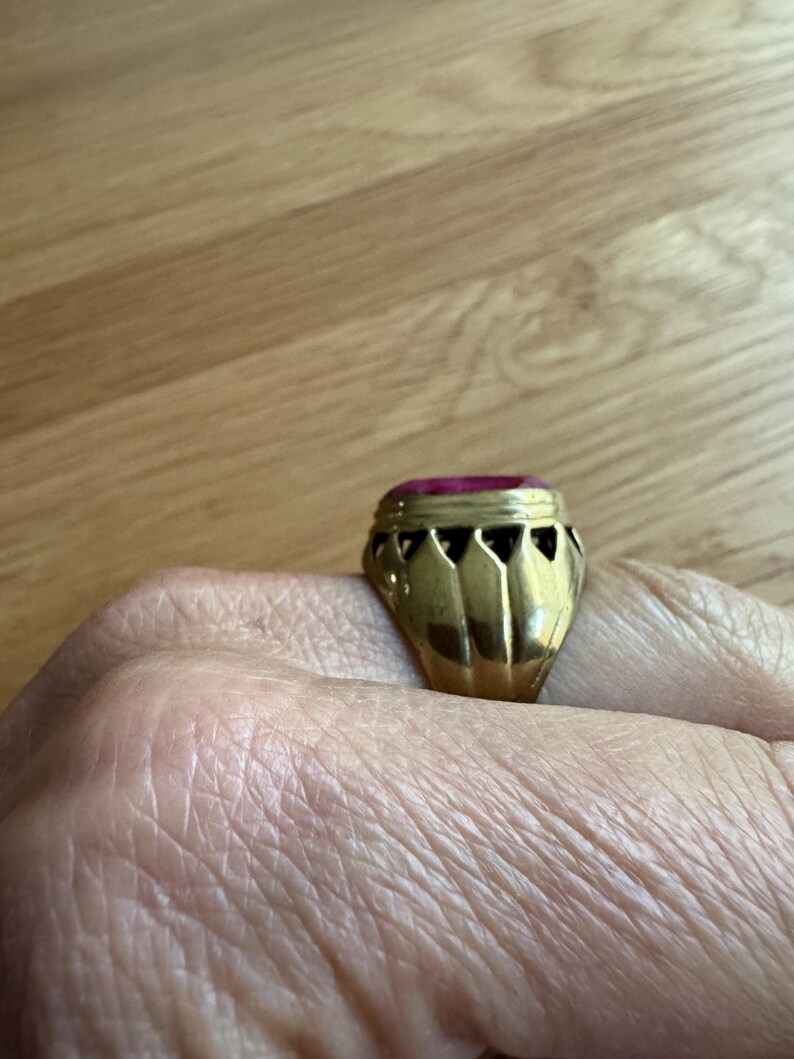 May include: A gold ring featuring a prominent, rectangular, deep pink gemstone set in a metal band. The setting displays a series of triangular cutouts along the gallery, creating a crown-like appearance around the base of the stone.