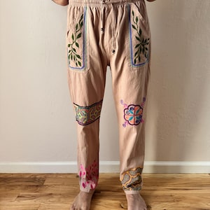 May include: Tan pants with embroidered floral and geometric designs. The pants have a drawstring waist and pockets with green leaf embroidery. Colorful embroidery accents the knees and ankles. The pants are made of a lightweight fabric.