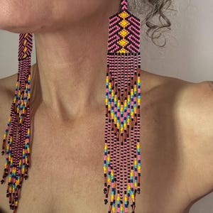 May include: Long, beaded earrings with a geometric design. The earrings feature a pink, black, yellow, blue, and brown color scheme. The top portion has a geometric pattern, and the bottom has long, dangling strands of beads.