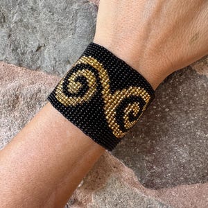 May include: Black beaded cuff bracelet with a gold spiral design. The bracelet is worn on a wrist and is made of small, tightly woven beads. The gold design is centered on the bracelet.