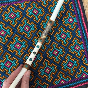 Peut inclure: Une flûte artisanale de couleur ivoire avec des bandes décoratives en cuir marron et en métal à motifs. La flûte présente des accents turquoise et est posée sur un textile brodé coloré avec un motif géométrique bleu, orange, rose et vert.