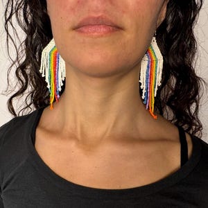 May include: A pair of rainbow beaded earrings with a white base. The earrings are long and dangle, with a fringe of beads at the bottom.