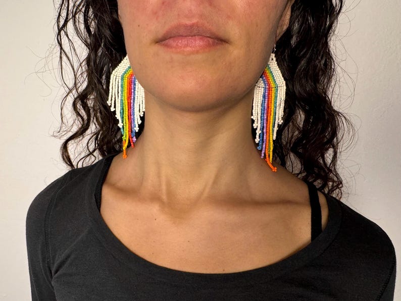 May include: A pair of rainbow beaded earrings with a white base. The earrings are long and dangle, with a fringe of beads at the bottom.