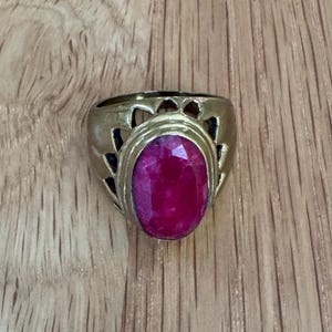 May include: A gold-toned metal ring featuring a large, oval-shaped, faceted magenta-pink gemstone set in the center. The band of the ring displays a decorative cutout pattern with triangular shapes surrounding the central stone setting.