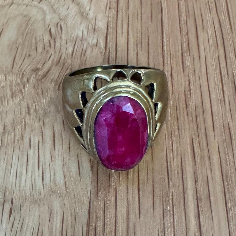 May include: A gold-toned metal ring featuring a large, oval-shaped, faceted magenta-pink gemstone set in the center. The band of the ring displays a decorative cutout pattern with triangular shapes surrounding the central stone setting.