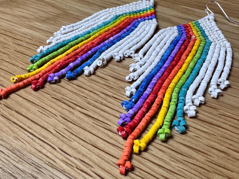 May include: A pair of rainbow-coloured beaded earrings with white beads. The earrings are long and dangle, with the beads arranged in a rainbow pattern.