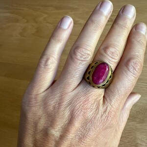 May include: A gold-toned metal ring featuring a large, oval-shaped, deep pink gemstone set in the center. The band of the ring has a wide, thick design with a series of triangular cutouts along the edges surrounding the central stone setting.
