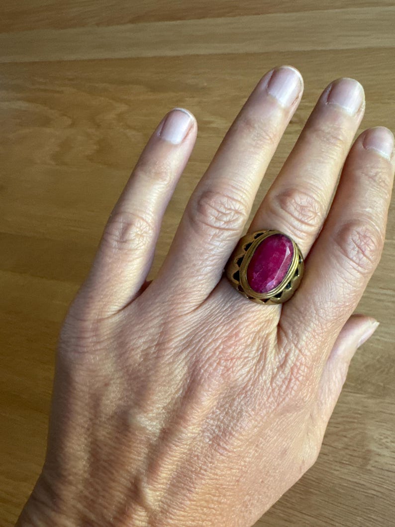 May include: A gold-toned metal ring featuring a large, oval-shaped, deep pink gemstone set in the center. The band of the ring has a wide, thick design with a series of triangular cutouts along the edges surrounding the central stone setting.