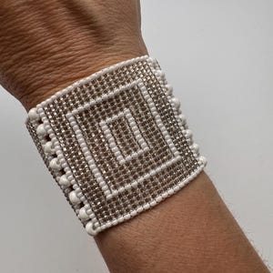 May include: A white beaded bracelet with a silver geometric design. The bracelet is made of small, round beads that are woven together to create a square pattern.