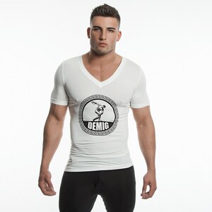 Pode incluir: Camiseta branca de gola em V com um gráfico preto e branco de uma pessoa lançando um disco. O gráfico é cercado por um design de chave grega preto e branco e a palavra "DEMIG" é impressa abaixo do gráfico.