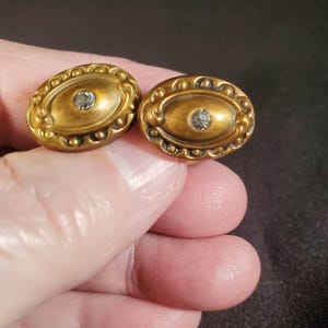 May include: A pair of oval-shaped brass-toned cufflinks featuring a raised decorative border with small bead-like accents and a single clear, faceted stone set in the center of each piece, showcasing a metallic finish and intricate detailing.