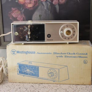 May include: A vintage Westinghouse automatic blanket clock control with a drowse alarm, featuring a white rectangular housing, a black clock face with silver hands, and a circular dial. The device sits atop its original cardboard box with blue text.