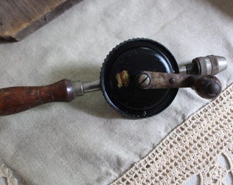 May include: An antique hand drill with a wooden handle and a black metal body. The drill has a crank handle and a chuck for holding drill bits. The tool is set against a neutral background.