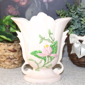 Puede incluir: Un jarrón de cerámica rosa pálido con un diseño floral. El jarrón tiene un borde festoneado y dos asas. Una flor rosa con hojas verdes está pintada en la parte delantera. El jarrón es un objeto decorativo.
