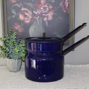 Peut inclure: Une casserole double fond en émail bleu marine profond avec un couvercle assorti et deux longues poignées fines s'étendant sur les côtés. La casserole présente une finition lisse et brillante ainsi qu'une forme cylindrique.