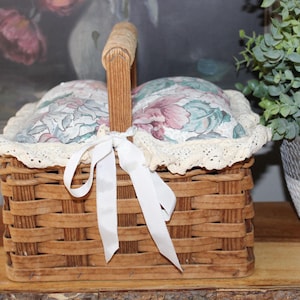 May include: A woven wicker basket with a white lace trim and a white satin ribbon. The basket is lined with two floral patterned cushions.