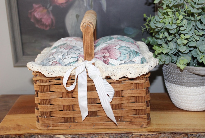 May include: A woven wicker basket with a white lace trim and a white satin ribbon. The basket is lined with two floral patterned cushions.