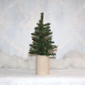 May include: A small, artificial evergreen tree in a beige stoneware crock. The tree has dark green needles and a conical shape. The crock is cylindrical with a slightly flared rim. The background is a light blue with a subtle pattern.