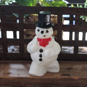 May include: A white snowman figurine with a black top hat, black button eyes, and a red scarf. The snowman has three black buttons down its front and is standing on a wooden surface. The background includes a decorative metal grate and greenery.