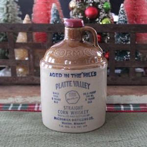 May include: A vintage ceramic whiskey jug with a brown top and handle, and a cream-colored body. The jug has blue text that reads "Aged in the Hills" and "Platte Valley Straight Corn Whiskey". The jug is 750 ml.