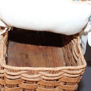 May include: A woven wicker basket with a white fabric lining and a white ribbon. The basket is empty and has a wooden base.