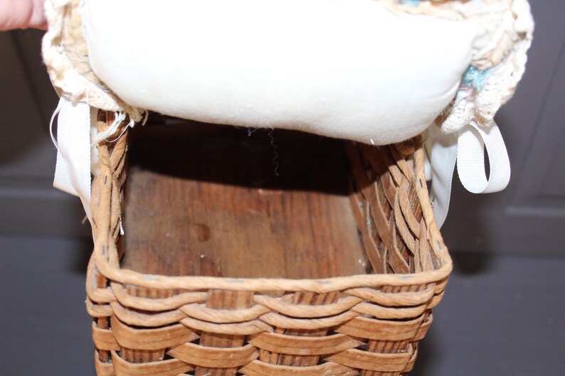 May include: A woven wicker basket with a white fabric lining and a white ribbon. The basket is empty and has a wooden base.
