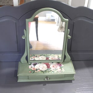 Op de afbeelding: Een saliegroene tafelspiegel met een boogvormige bovenkant, voorzien van een enkele lade met een heldere kristallen knop. De basis en het frame zijn versierd met een bloemenprint van roze, witte en donkerrode rozen en groen blad.